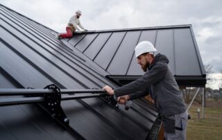 Two workers in hard hats install black metal roofing on a steeply pitched roof. The sky is overcast.