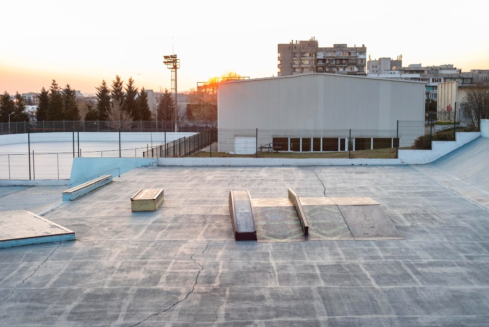 Empty skate park at sunset with various ramps and rails. A fenced sports court and buildings are in the background.